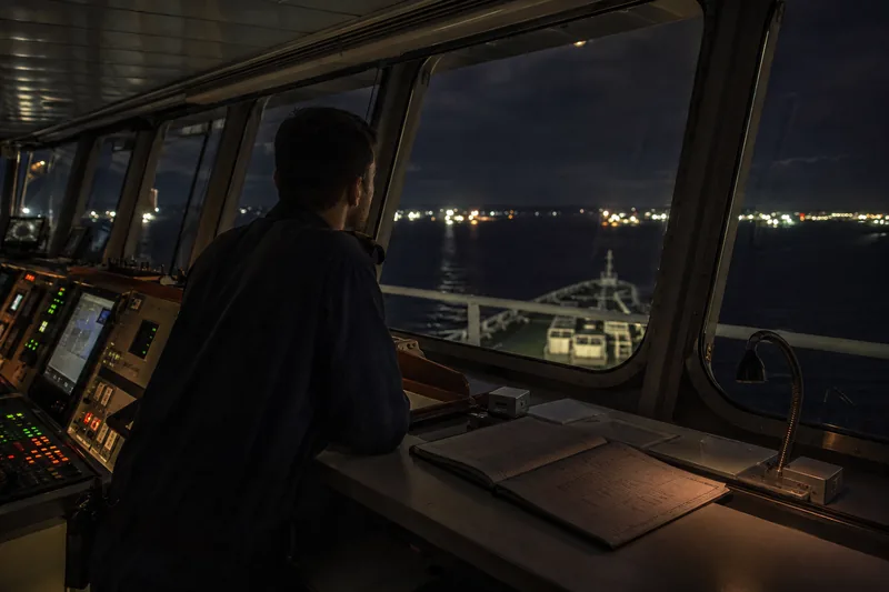 A crew member standing watch on a ship’s bridge at night, a logbook open on the desk beside them.